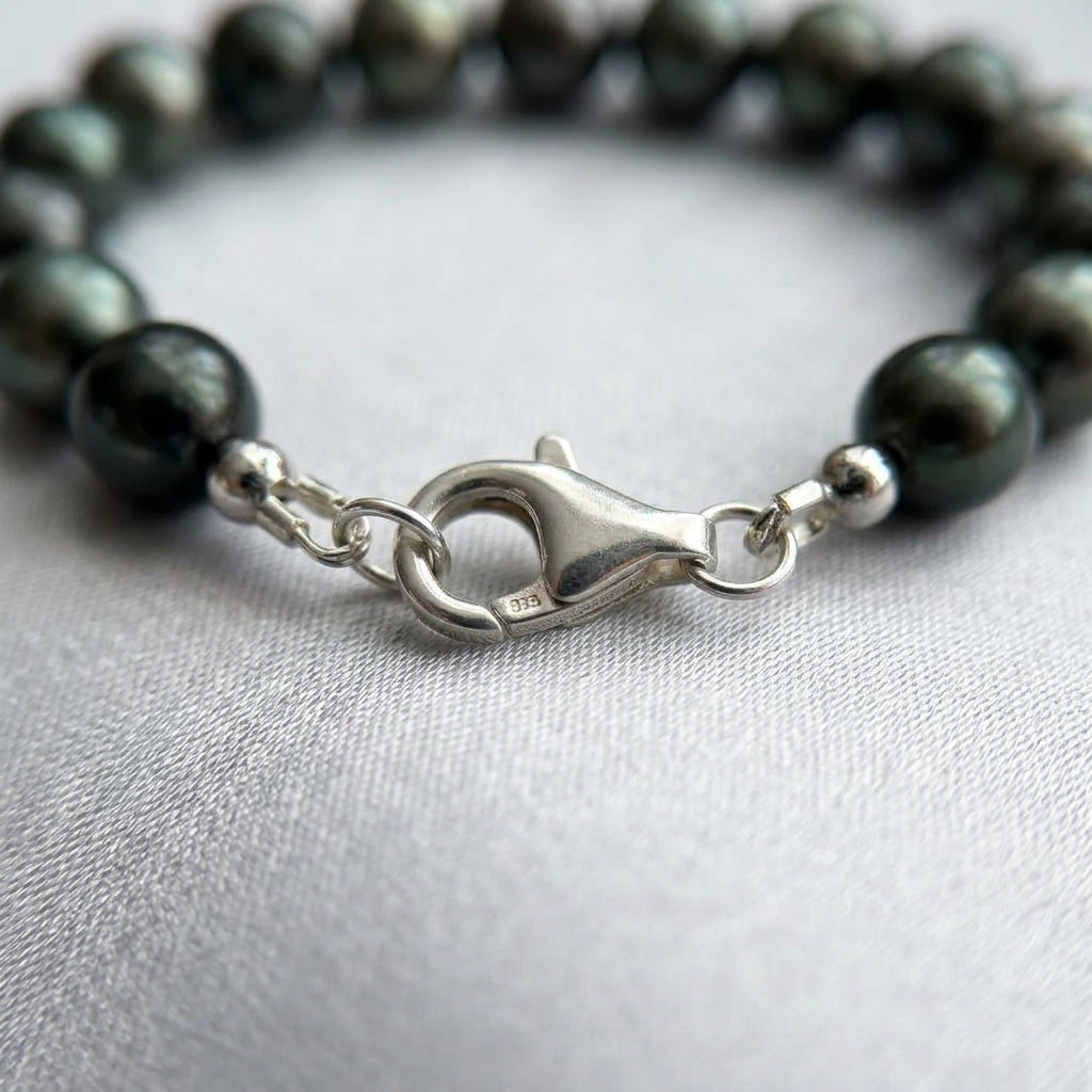 Close-up of a fastened sterling silver lobster clasp stamped 925, joining dark peacock baroque pearls with green-teal reflections on pale satin.⠋