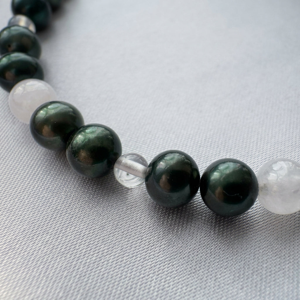 Close-up of dark peacock baroque pearls with green-teal reflections, interspersed with a clear quartz bead and milky white moonstone beads on pale satin.