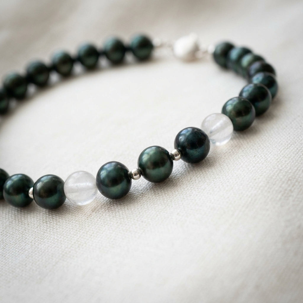 Peacock baroque pearl bracelet arranged in an oval on cream fabric, showing green-teal lustre, pale translucent gemstone accents and small silver-coloured spacers, with the clasp softly blurred in the background.