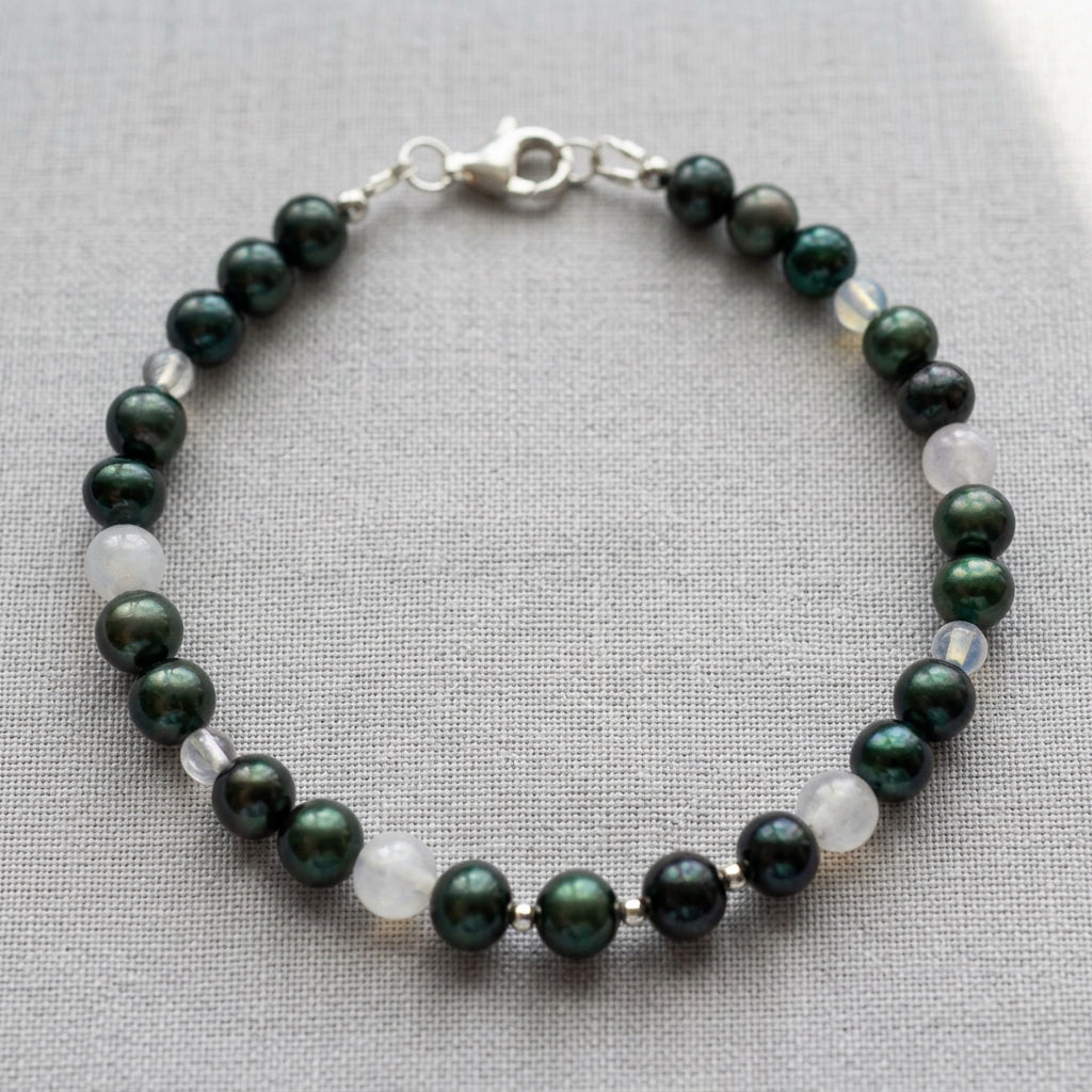 Peacock baroque pearl bracelet laid in an oval on grey linen, with milky moonstone and clear quartz beads, small silver-coloured spacers and a fastened sterling silver lobster clasp.