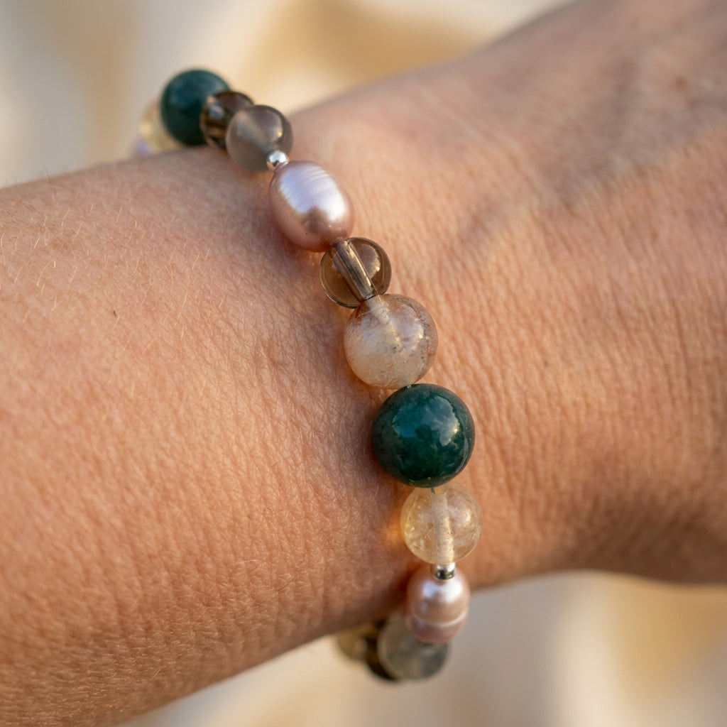 Angled close-up of a pink baroque pearl and gemstone bracelet worn on a wrist in warm light, showing mottled green, translucent smoky brown, pale grey and honey-yellow beads with small sterling silver spacers.