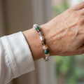 Mauve baroque pearl bracelet with teal-green, smoky brown, pale grey and golden translucent gemstone beads, worn on the wrist beside a white silk shirt cuff.
