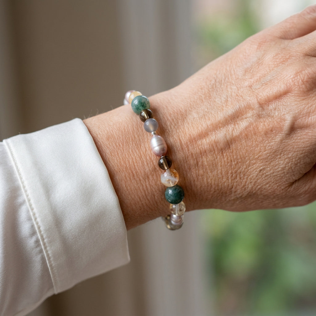 Mauve baroque pearl bracelet with teal-green, smoky brown, pale grey and golden translucent gemstone beads, worn on the wrist beside a white silk shirt cuff.