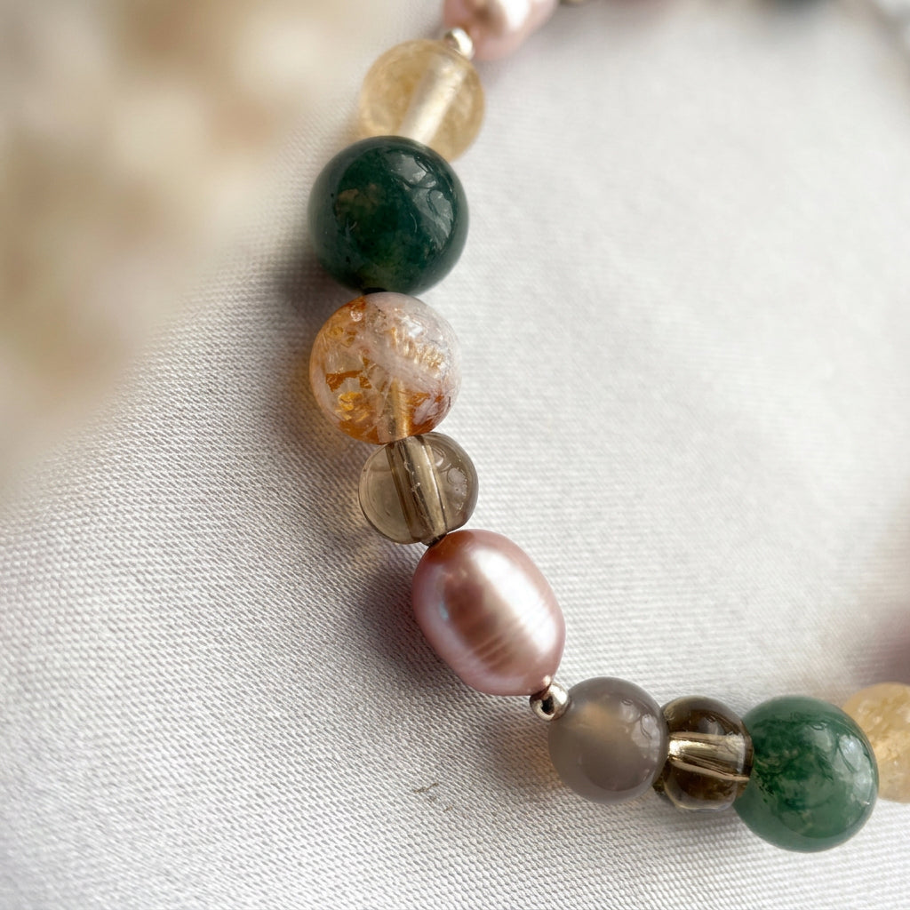Close-up of a lustrous mauve-pink baroque pearl among smoky brown, pale grey, mottled green and golden-yellow gemstone beads, arranged in a curve on white fabric.