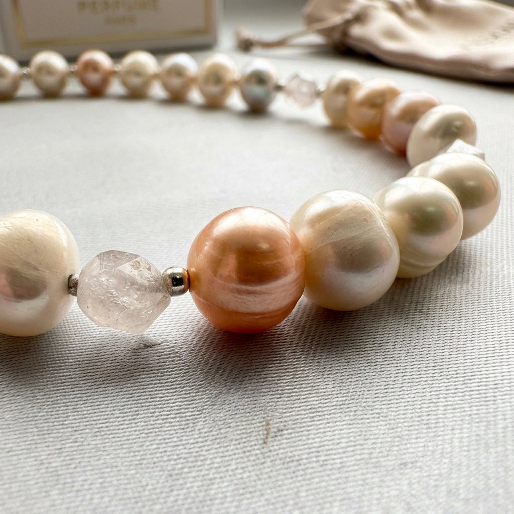 Close-up of creamy white and peach baroque pearls beside a faceted clear quartz bead flanked by sterling silver spacers, arranged in a curve on pale fabric.