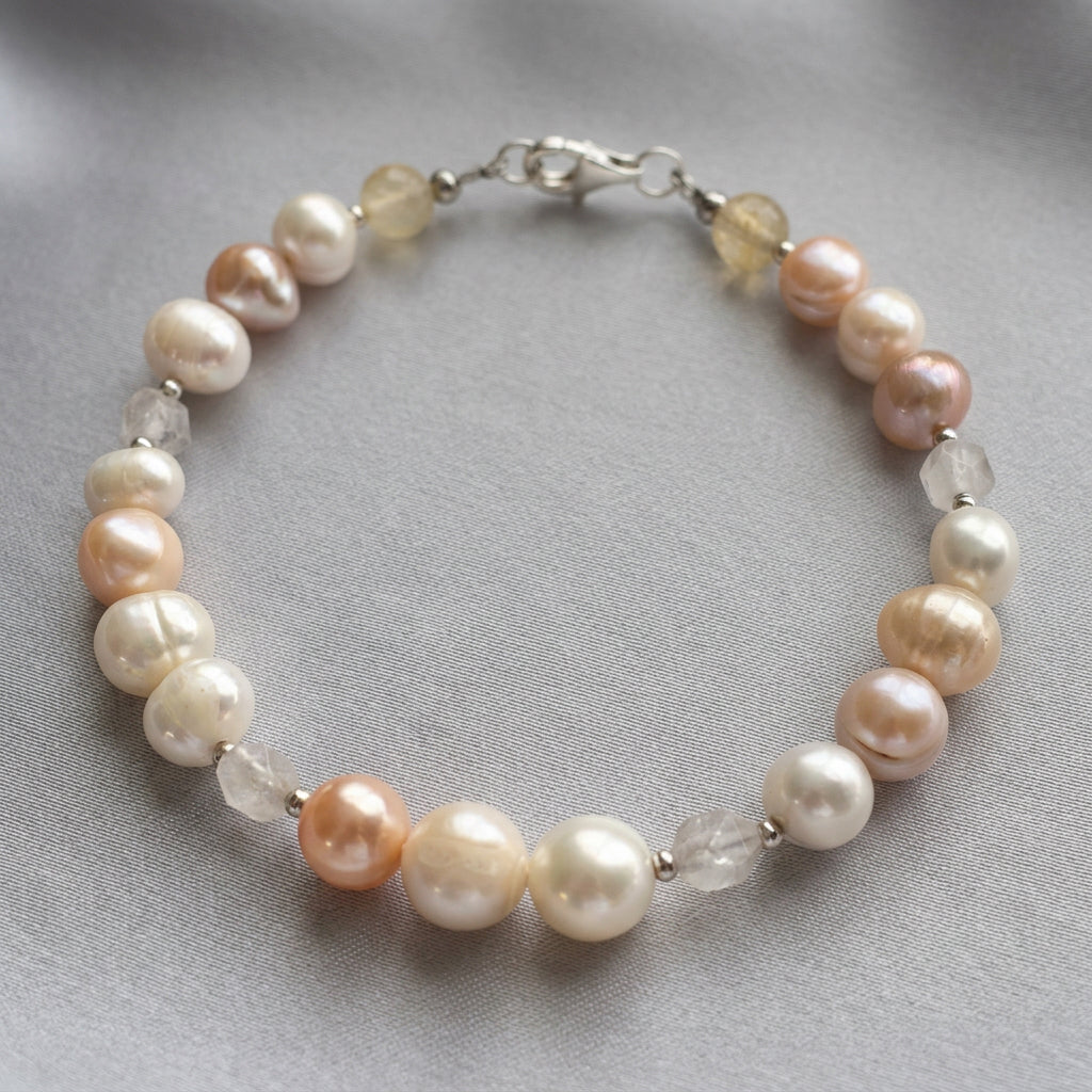 Baroque pearl bracelet arranged in an oval on grey fabric, showing creamy white and peach pearls, faceted clear quartz, two pale yellow citrine beads and a sterling silver lobster clasp.