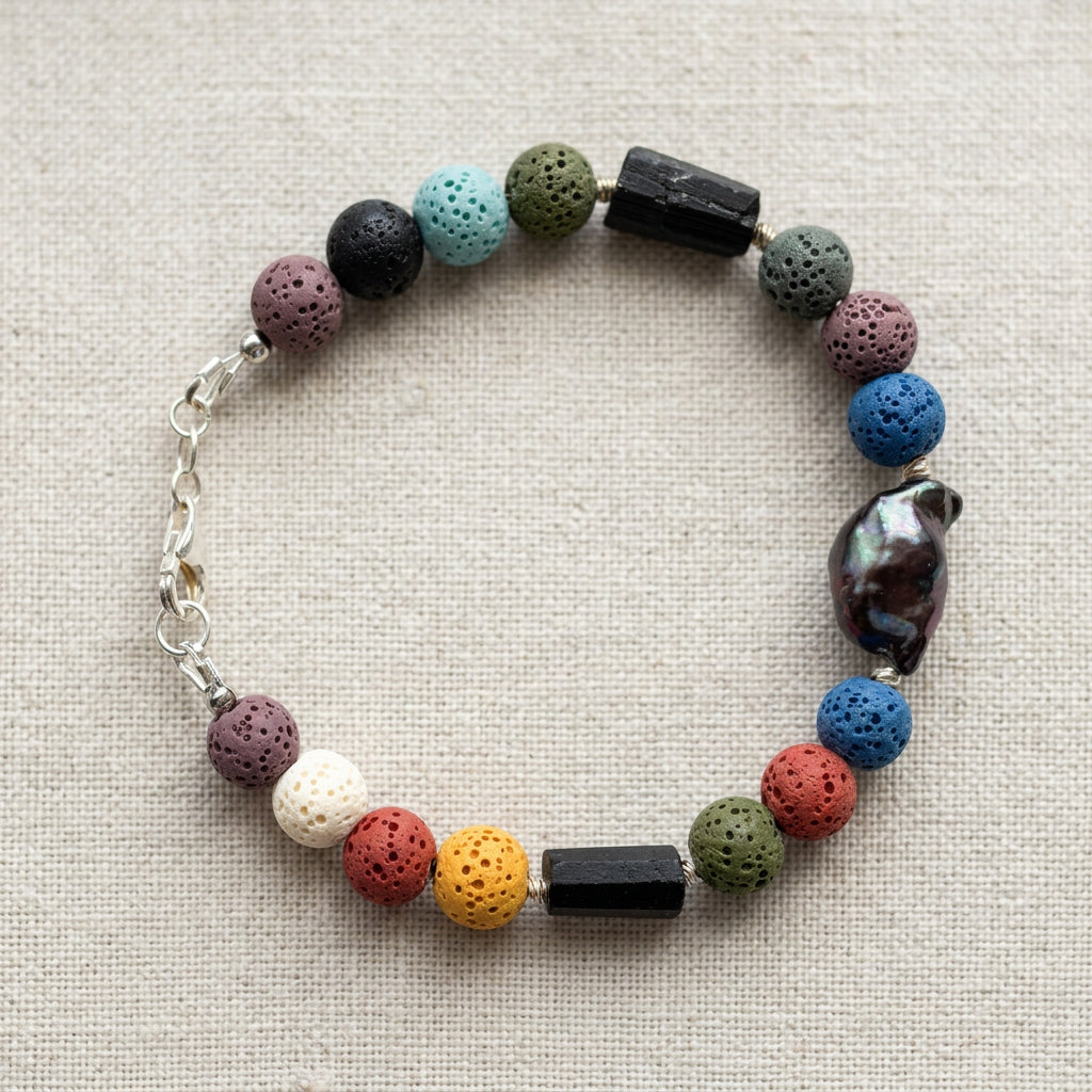 Dark blue keshi pearl bracelet arranged in a circle on cream linen, with multicoloured porous lava beads, two raw black tourmaline pieces, sterling silver spacers and a fastened lobster clasp.