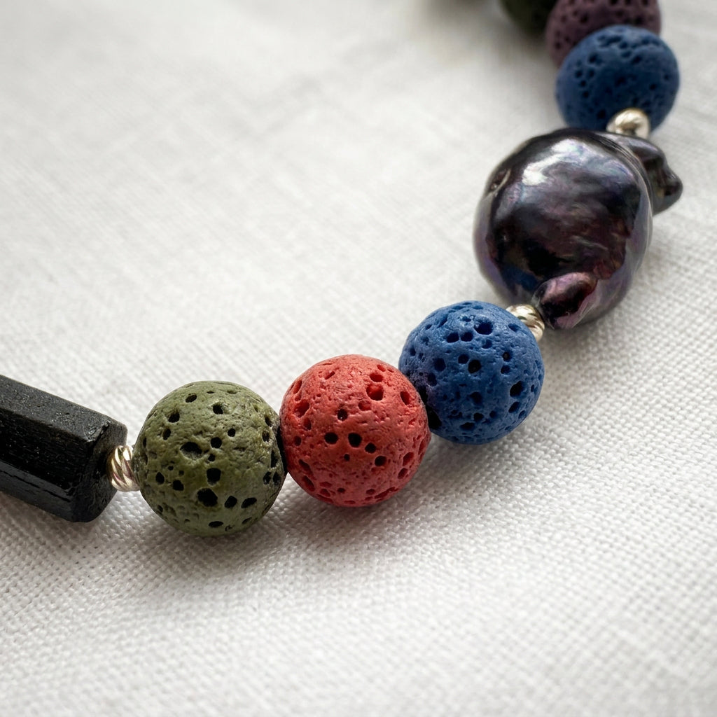 Close-up of porous olive-green, coral-pink and blue lava beads beside a lustrous dark blue keshi pearl, raw black tourmaline and sterling silver spacers on cream linen.
