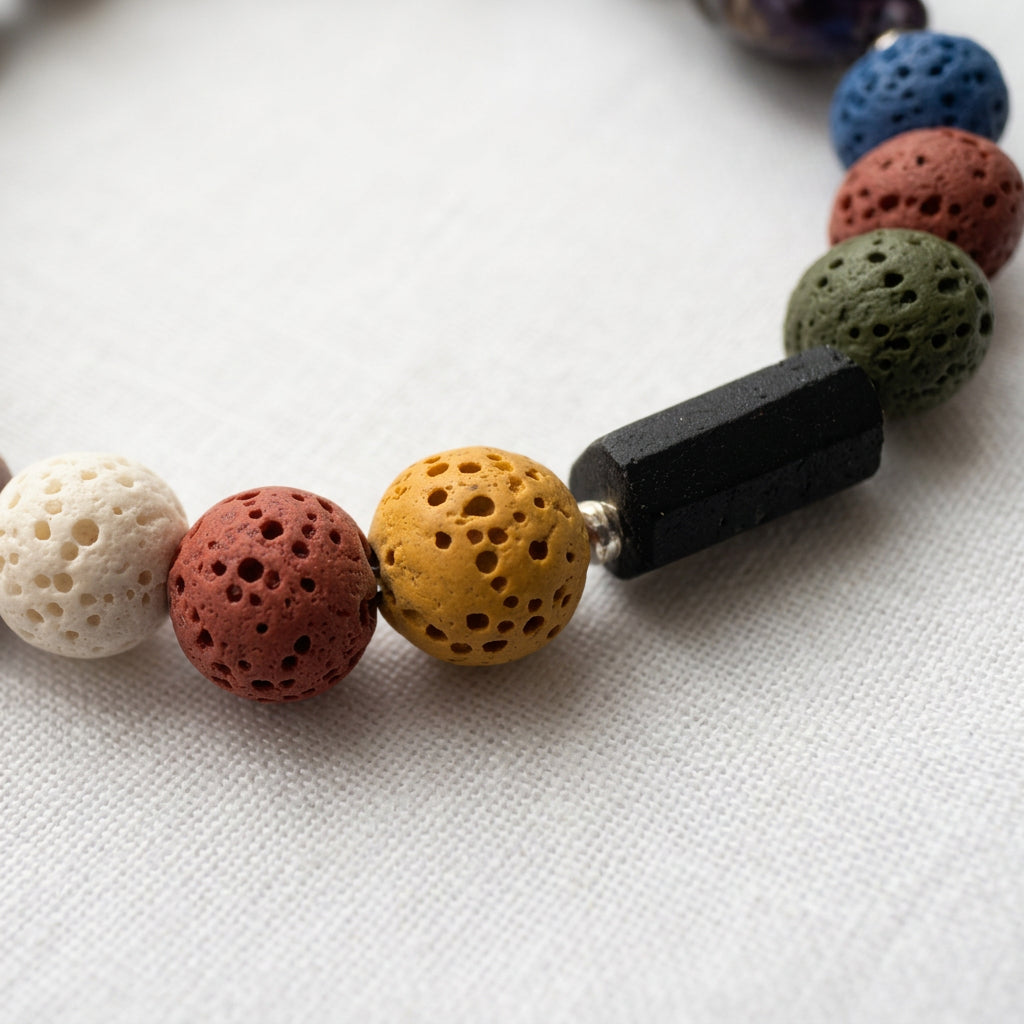 Close-up of raw black tourmaline beside porous mustard-yellow, coral-pink, cream and olive-green lava beads, with sterling silver spacers on white linen.
