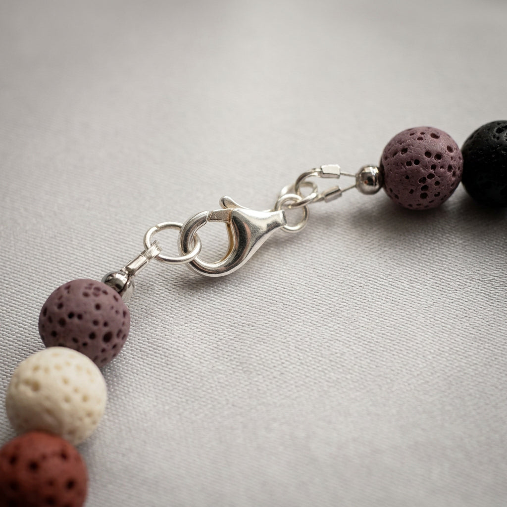 Close-up of a fastened sterling silver lobster clasp between porous mauve lava beads, with cream, coral-pink and black lava beads on pale fabric.