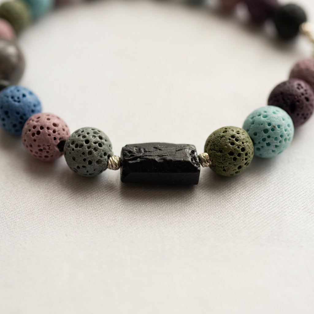Close-up of a raw black tourmaline piece framed by twisted sterling silver spacers and porous grey-green, olive, turquoise and mauve lava beads on cream fabric.