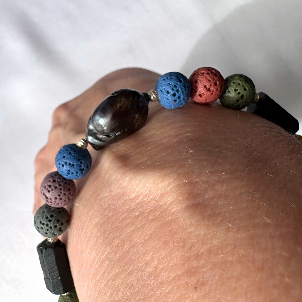 Sunlit close-up of a dark blue keshi pearl bracelet worn on a wrist, showing the pearl’s metallic lustre, colourful porous lava beads, raw black tourmaline and sterling silver spacers.