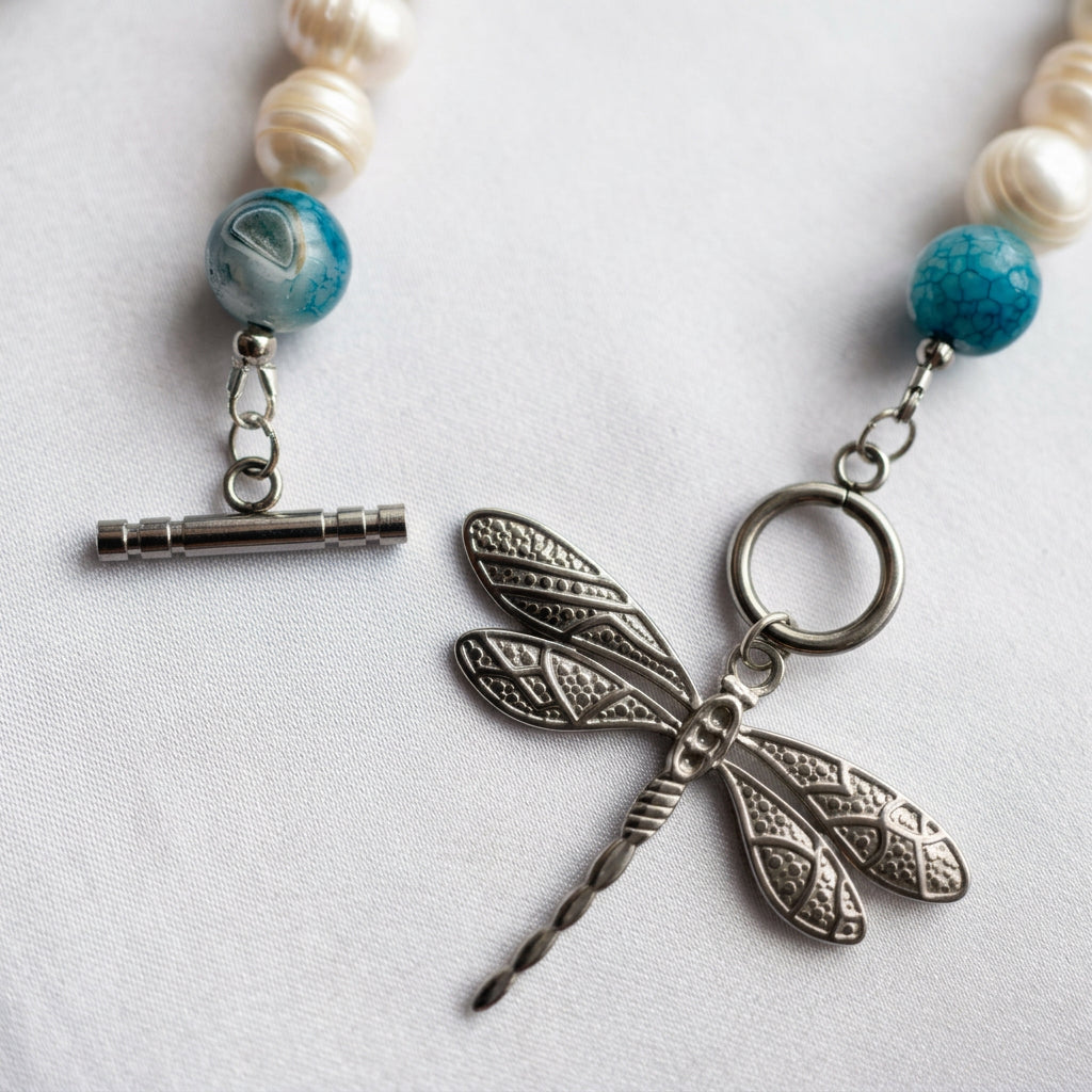 Close-up of an unfastened silver-coloured toggle clasp, showing the separate bar and ring beside a detailed stainless steel dragonfly pendant, blue glass beads and white baroque pearls on pale fabric.⠋
