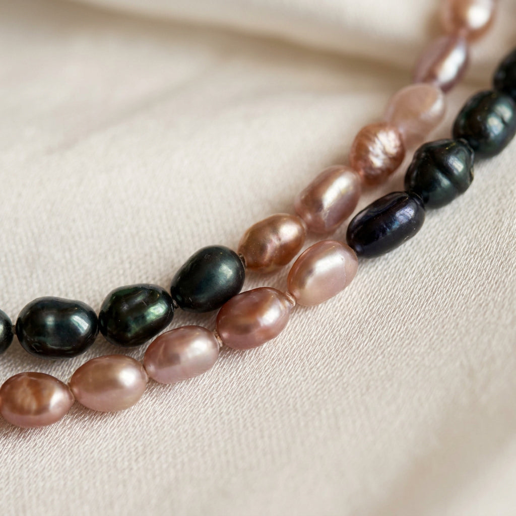 Close-up of blush-pink and dark blue rice pearl strands on cream satin, showing their irregular oval shapes, natural surface ridges and pink, green and blue reflections.