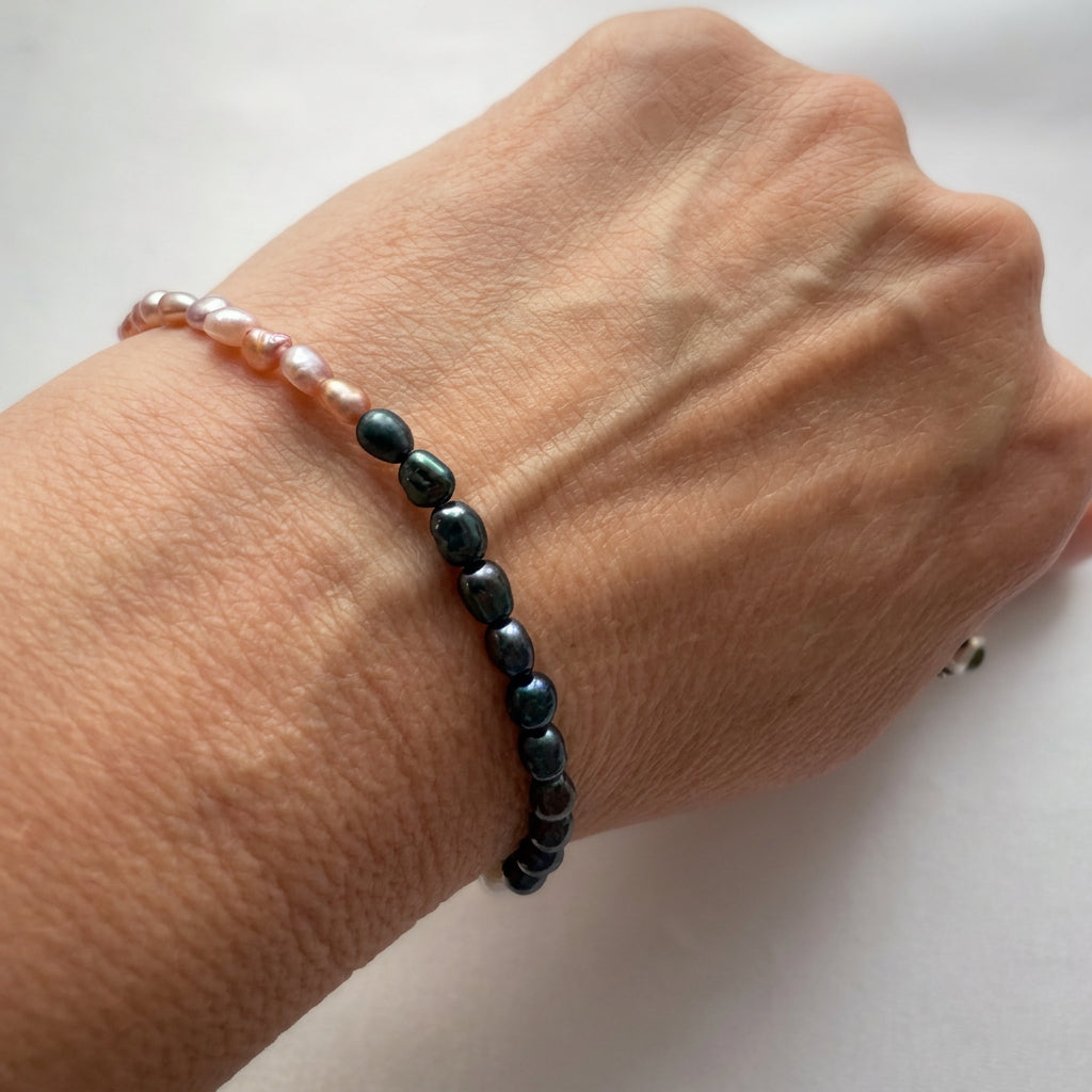 Rice pearl bracelet worn on a wrist, showing a section of lustrous dark blue pearls meeting blush-pink pearls, with naturally irregular shapes and green-blue reflections.