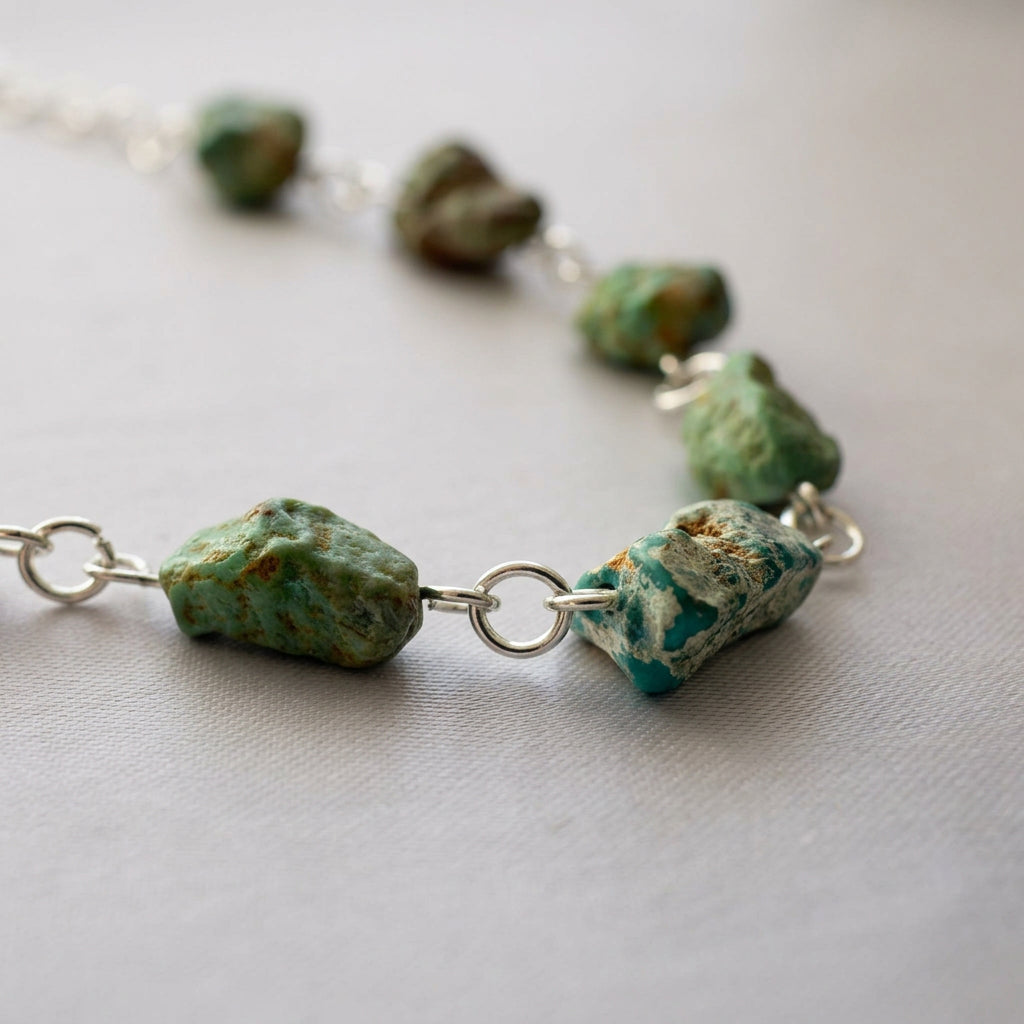 Close-up of raw green-blue Hubei turquoise stones with textured brown and cream matrix, joined by sterling silver rings in a curve on cream fabric.