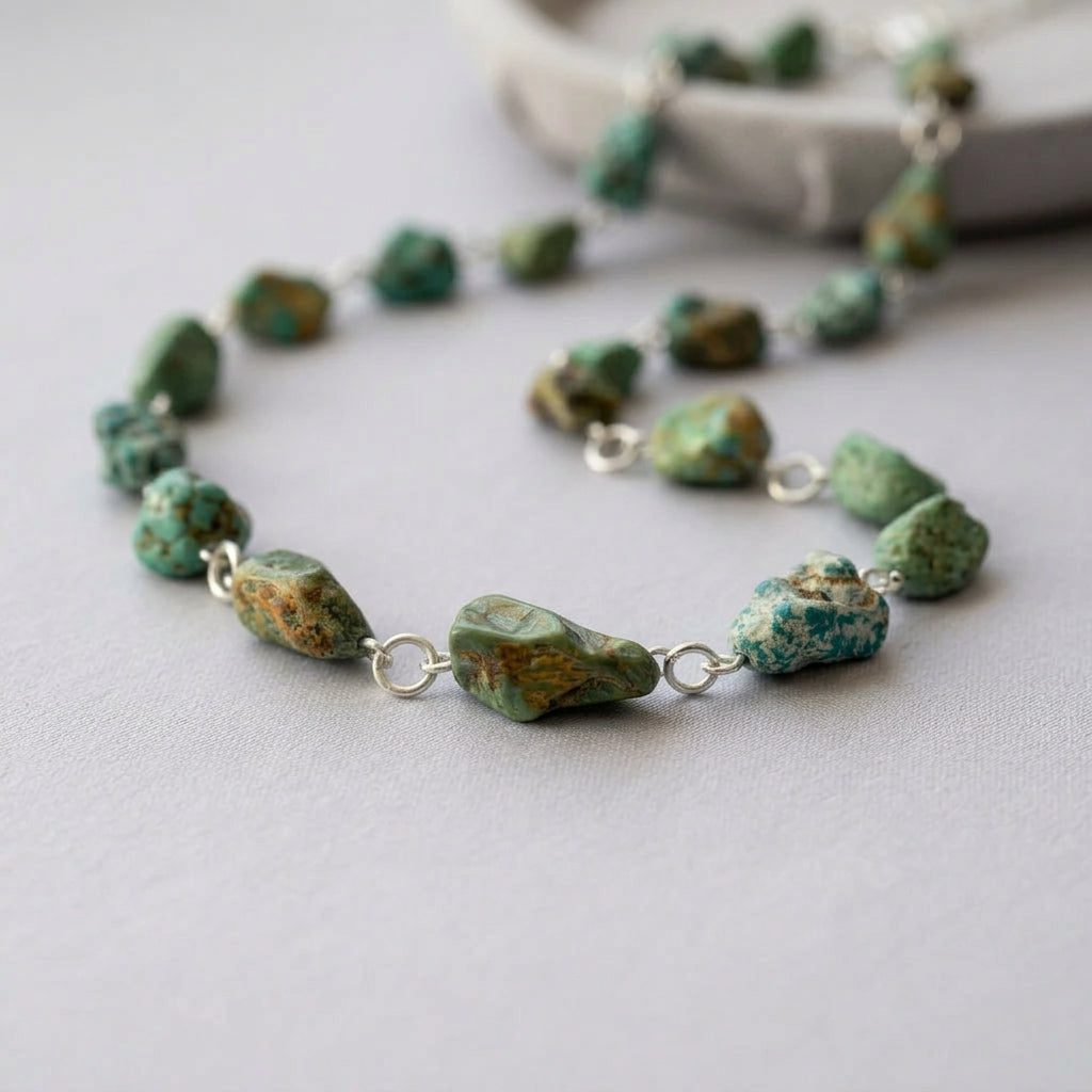 Raw Hubei turquoise necklace loosely curved on pale fabric, showing irregular green-blue stones with rough brown matrix and handmade sterling silver wire links, with a white dish softly blurred behind.
