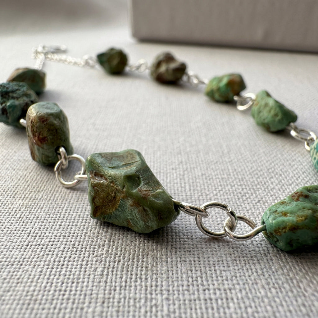 Close-up of an angular raw green Hubei turquoise stone with brown matrix and natural surface hollows, joined to neighbouring stones by handmade sterling silver wire loops on pale linen.