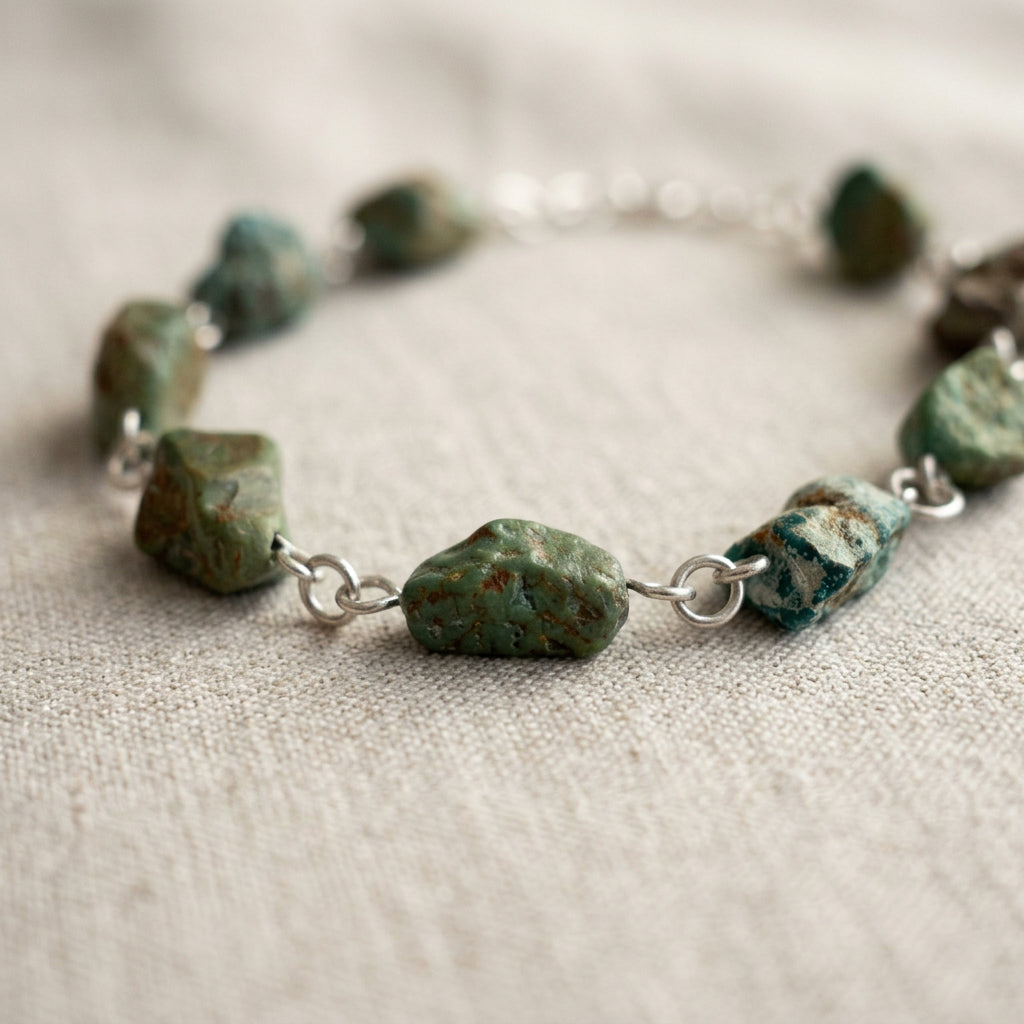 Close-up of raw green-blue Hubei turquoise stones with rough brown and cream matrix, joined by handmade sterling silver wire loops on pale linen, with the necklace curving softly out of focus behind.