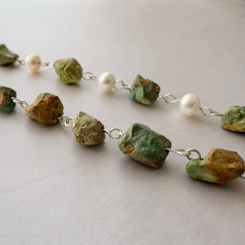 Close-up of raw green-blue Hubei turquoise stones with rough ochre-brown matrix, linked with sterling silver wire on cream satin, with three white baroque pearls along the strand behind them.