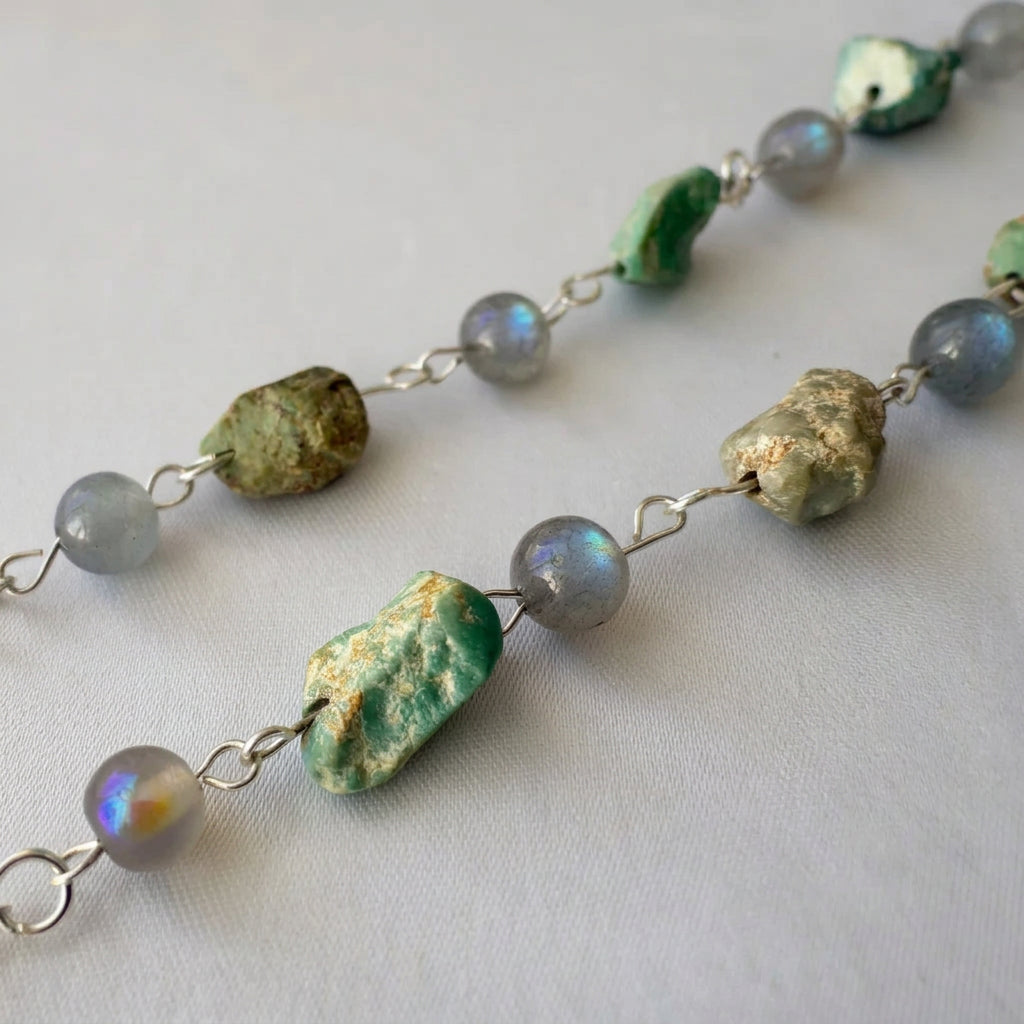 Close-up of raw green Hubei turquoise stones with earthy matrix and round labradorite beads flashing blue and gold, linked with sterling silver on cream fabric.
