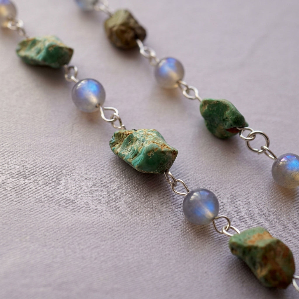 Close-up of textured green Hubei turquoise stones alternating with round labradorite beads showing vivid blue flashes, joined by sterling silver links on cream fabric.