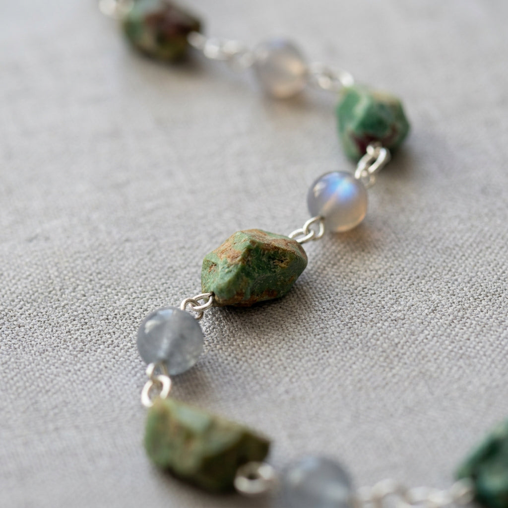 Close-up of a raw green Hubei turquoise stone with brown markings between round grey labradorite beads, one flashing blue, linked in sterling silver on cream linen.