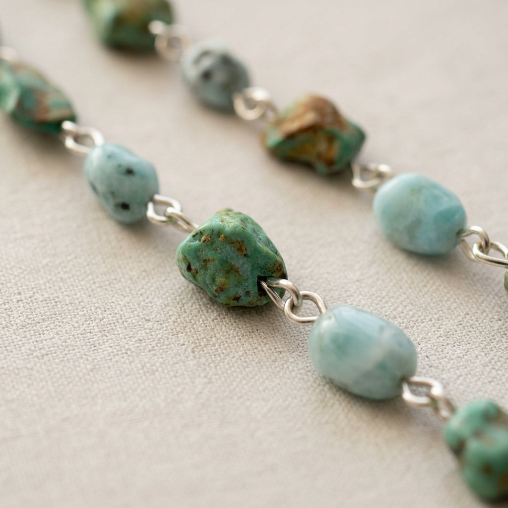 Close-up of raw green Hubei turquoise stones with brown matrix alternating with pale blue larimar beads, joined by sterling silver links on cream fabric.