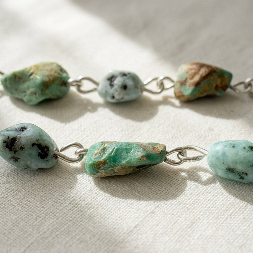 Close-up of raw green Hubei turquoise stones with brown matrix and pale blue larimar beads with dark markings, linked in sterling silver on sunlit cream linen.