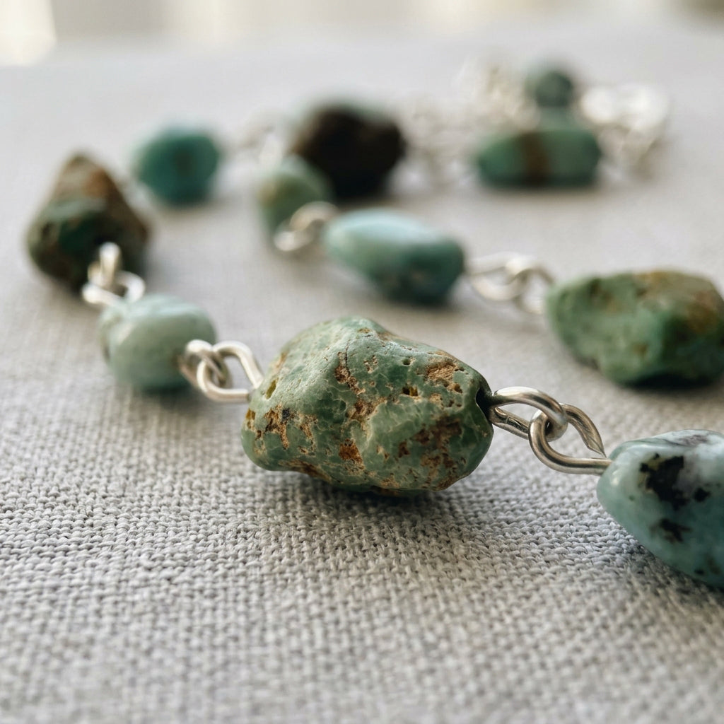 Close-up of a textured raw green Hubei turquoise stone with brown matrix, linked in sterling silver to pale blue larimar beads on cream linen.⠋