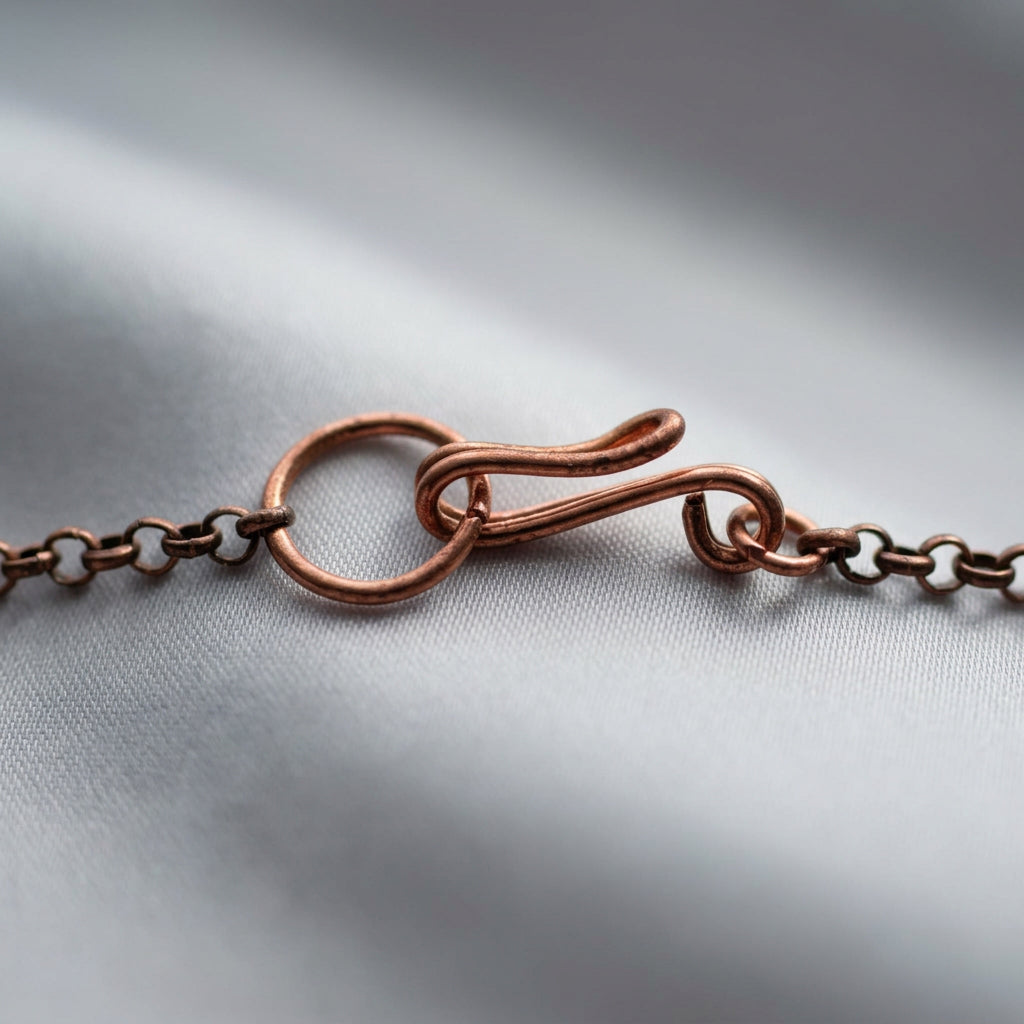 Close-up of a handmade copper wire hook clasp fastened through a circular ring, with copper chain links resting on soft grey fabric.