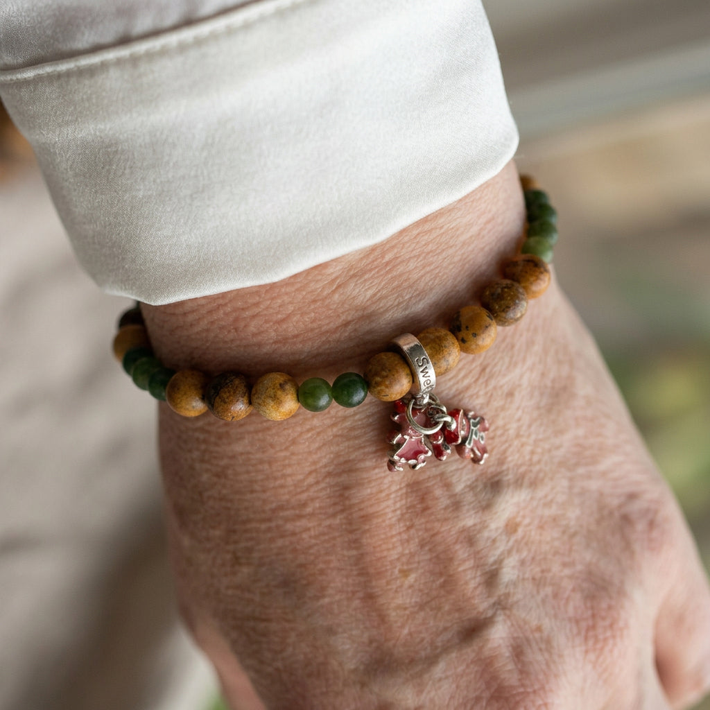 Mottled tan picture jasper and green jade bead bracelet with festive enamel gingerbread and candy cane charms, worn on the wrist beside a white silk shirt cuff.