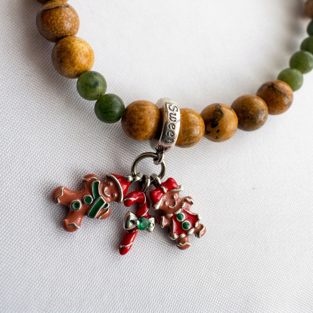 Close-up of two enamel gingerbread figures and a red candy cane charm beside picture jasper and green jade beads on white fabric.