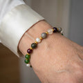 Mixed bead bracelet with green, burgundy, mottled grey and translucent stones, white pearls and gold-coloured spacers, worn on the wrist beside a white silk shirt cuff.