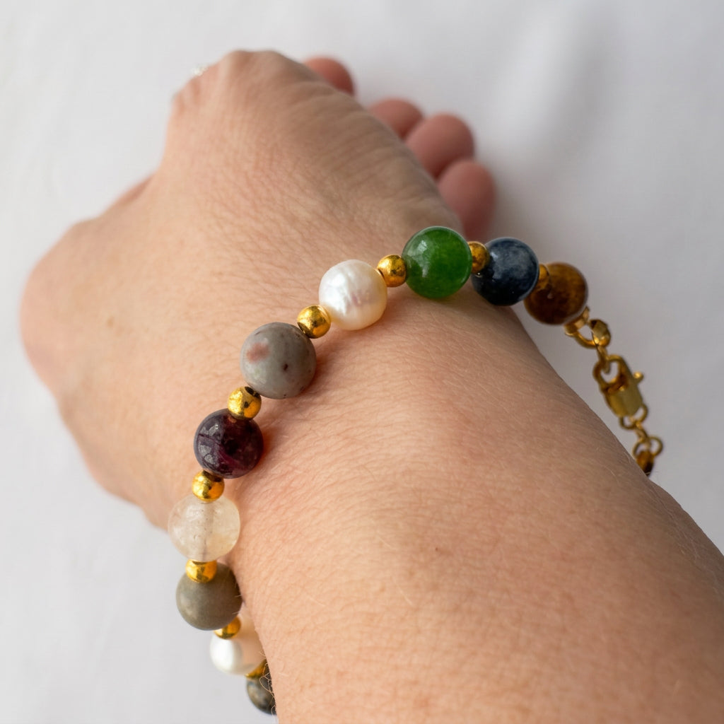 Side view of a beaded bracelet worn on a wrist, showing green, burgundy, grey and translucent stones, white baroque pearls, gold-coloured spacers and a gold-filled lobster clasp.