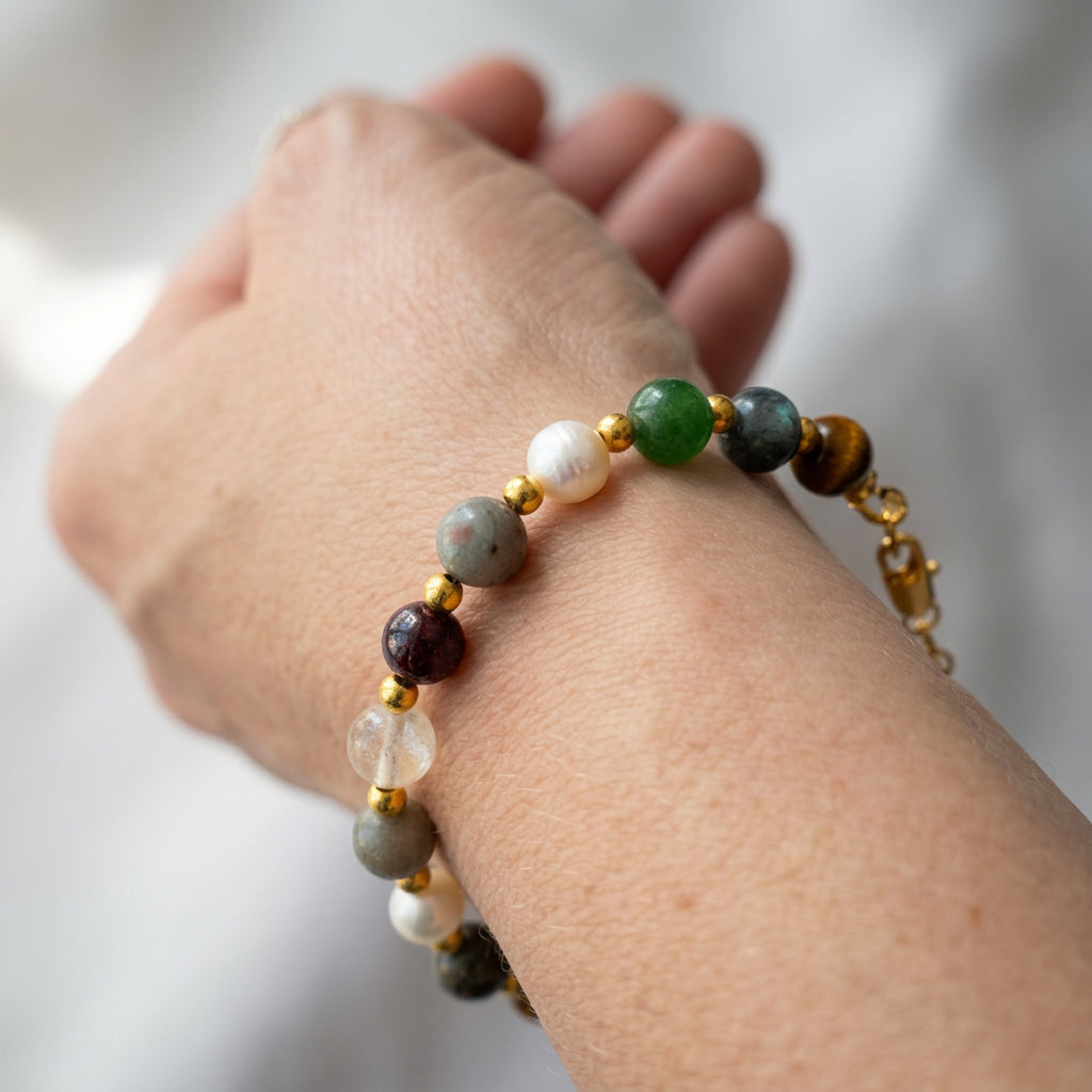 Beaded bracelet worn on a wrist, showing green, deep red, grey, brown and translucent stones with white baroque pearls, gold-coloured spacers and a gold-filled lobster clasp.⠋