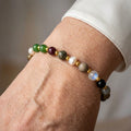 Mixed gemstone and white baroque pearl bracelet with green, burgundy, mottled grey and translucent beads separated by gold-coloured spacers, worn beside a white silk shirt cuff.