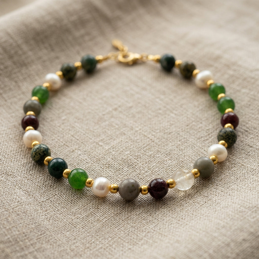 Beaded bracelet arranged in an oval on beige linen, featuring green, burgundy, grey and translucent stones, white baroque pearls, gold-coloured spacers and a lobster clasp.