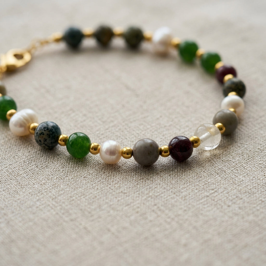 Beaded bracelet on cream linen, showing green jade, deep red garnet, grey jasper, a translucent bead and white baroque pearls separated by gold-coloured spacers.