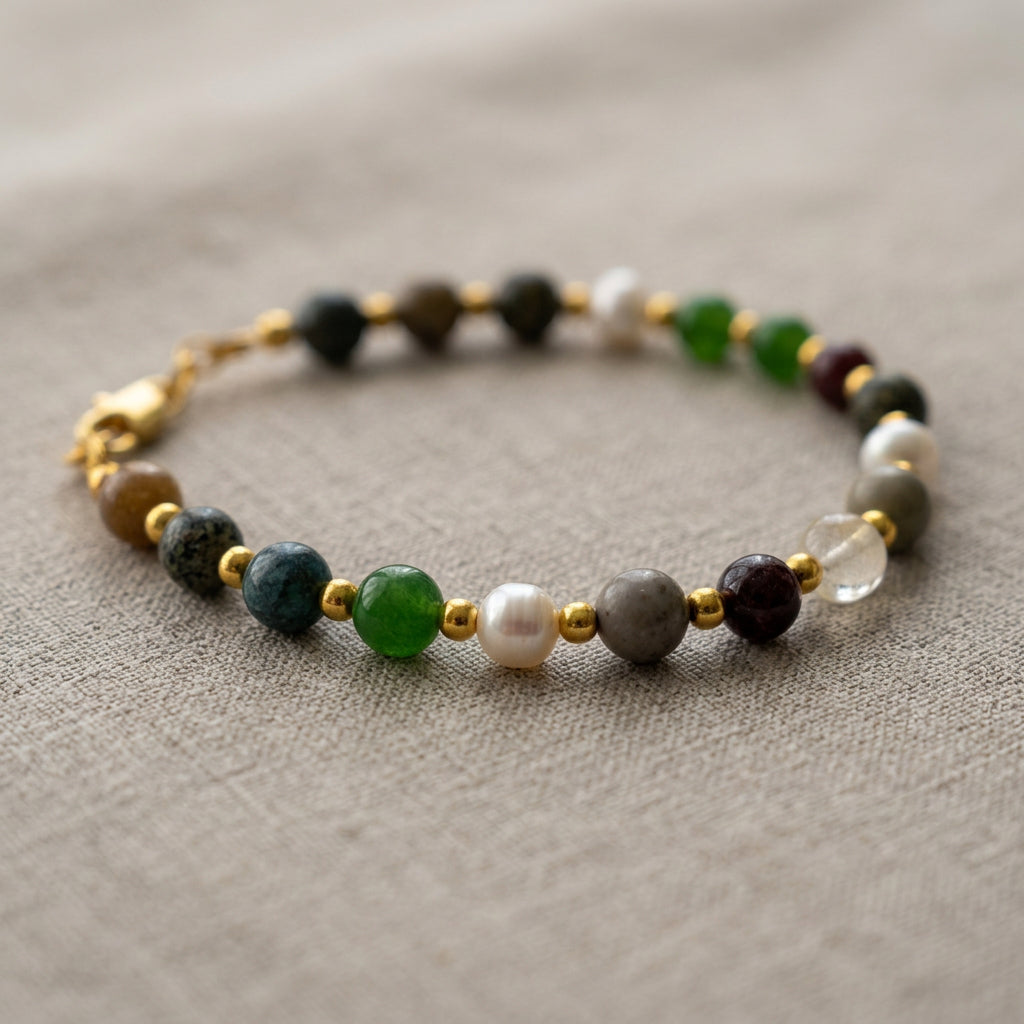 Beaded bracelet on beige linen, with green, grey, deep burgundy and translucent beads alongside white baroque pearls, separated by small gold-coloured spacers.