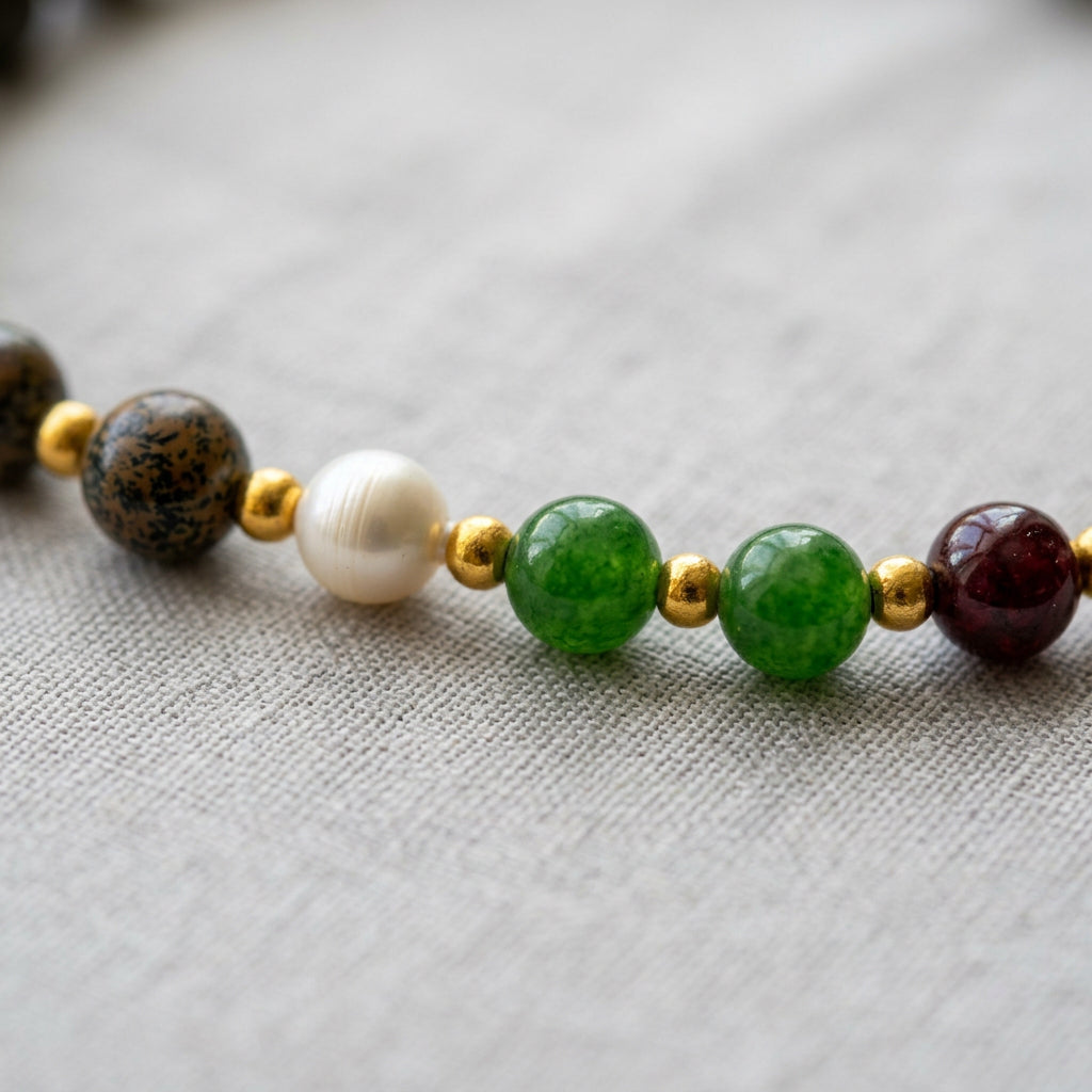 Close-up of two green beads beside a white baroque pearl, a patterned brown-grey bead and a deep burgundy bead, separated by gold-coloured spacers on pale linen.