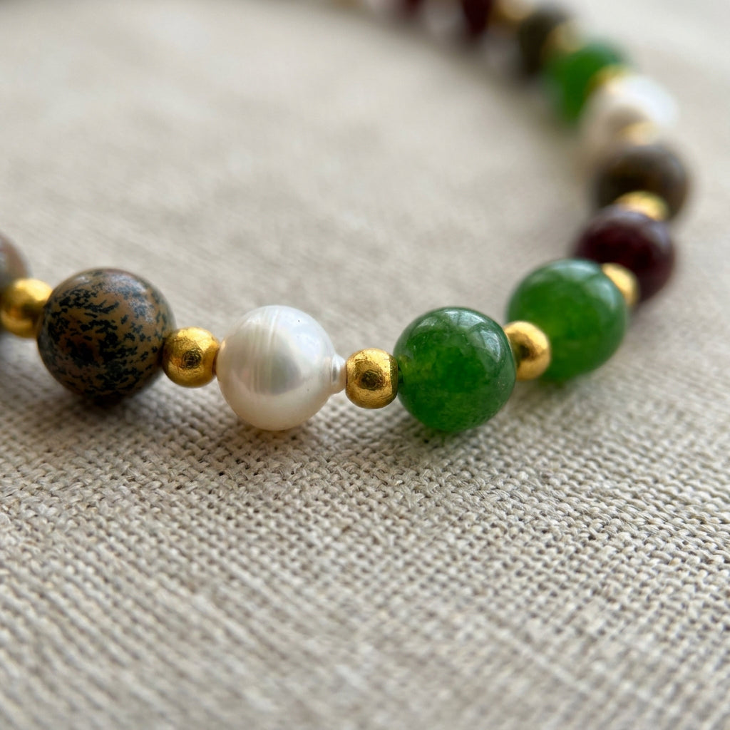 Close-up of green jade beads, a white baroque pearl and a patterned jasper bead separated by gold-coloured spacers on a bracelet resting on cream linen.