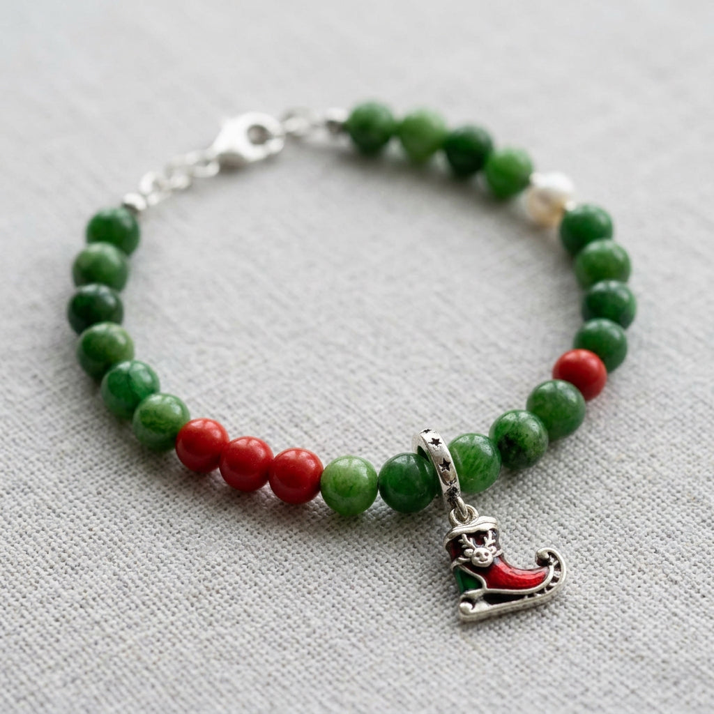 Green jade bracelet with red coral accents, a white pearl and a red enamel Santa’s sleigh charm, arranged on grey linen.