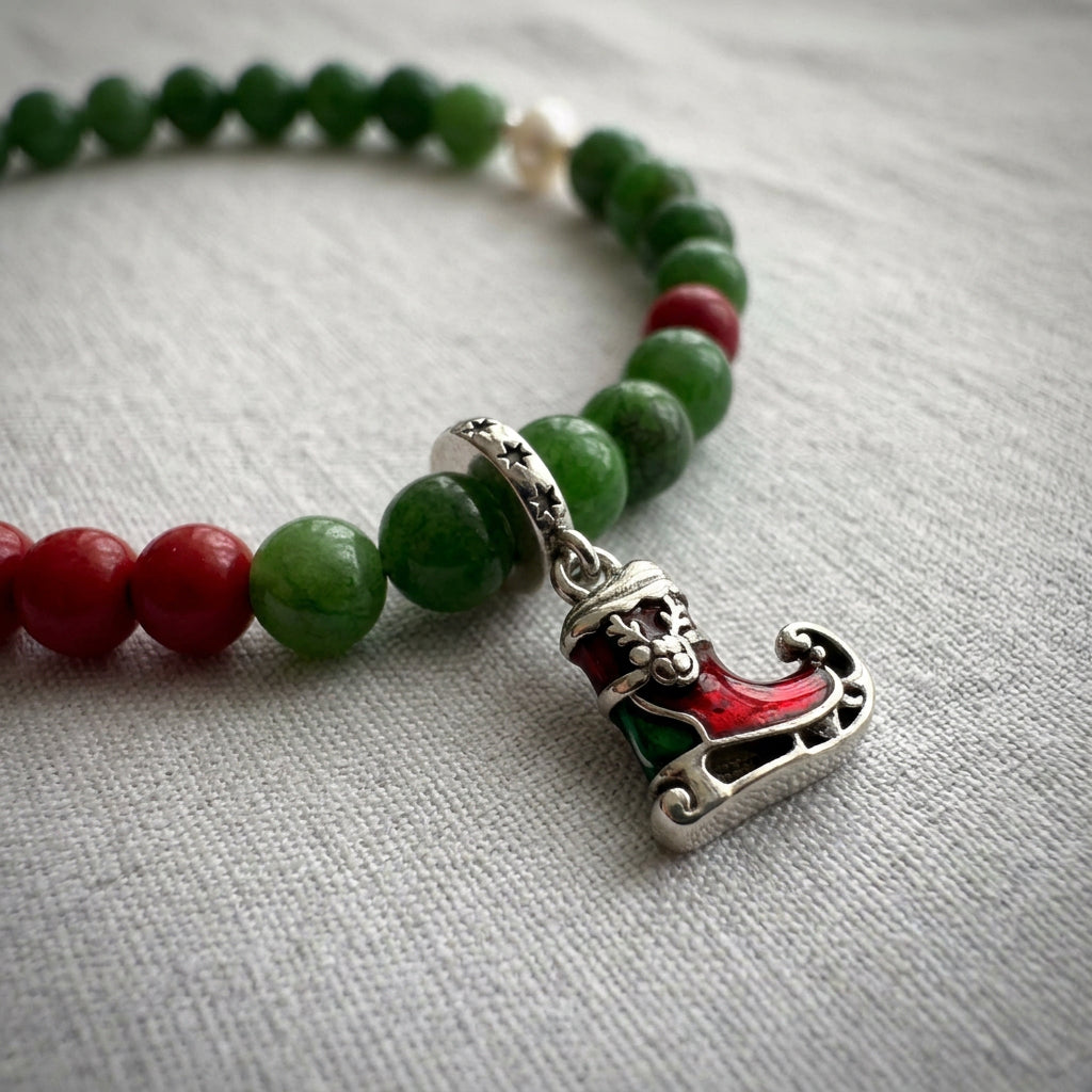 Close-up of a red and green enamel sleigh charm with a reindeer motif beside green jade and red coral beads on grey linen.