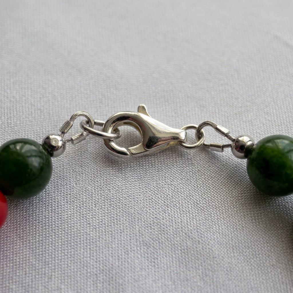 Close-up of the silver-coloured lobster clasp and connecting rings between green obsidian beads on white fabric.