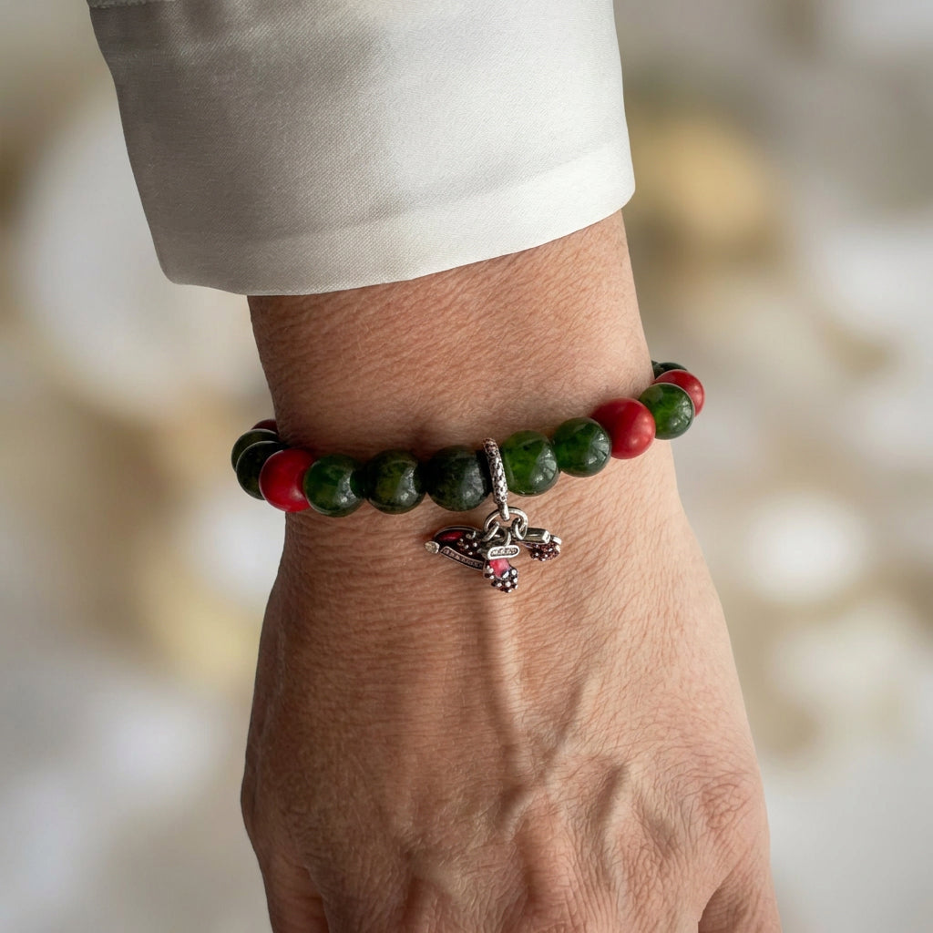Deep green obsidian bracelet with red coral beads and dangling Santa hat and stocking charms, worn on the wrist beside a white silk shirt cuff.