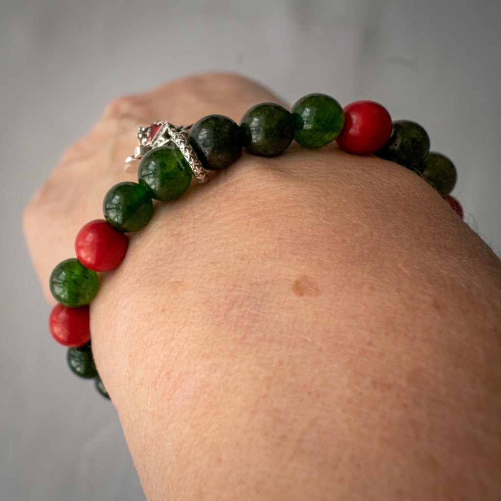 Angled wrist view of a green obsidian and red coral bracelet, with translucent green beads and Christmas charms to one side.
