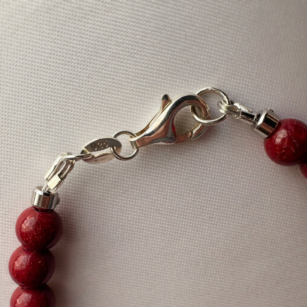 Close-up of the silver-coloured lobster clasp and connecting rings on a red coral bracelet, resting on cream fabric.⠋