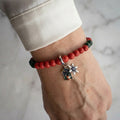 Red coral bracelet with deep green sandstone accents, a silver-coloured snowflake and colourful enamel advent window charm, worn on the wrist beside a white silk shirt cuff.