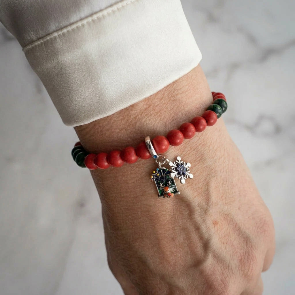 Red coral bracelet with deep green sandstone accents, a silver-coloured snowflake and colourful enamel advent window charm, worn on the wrist beside a white silk shirt cuff.